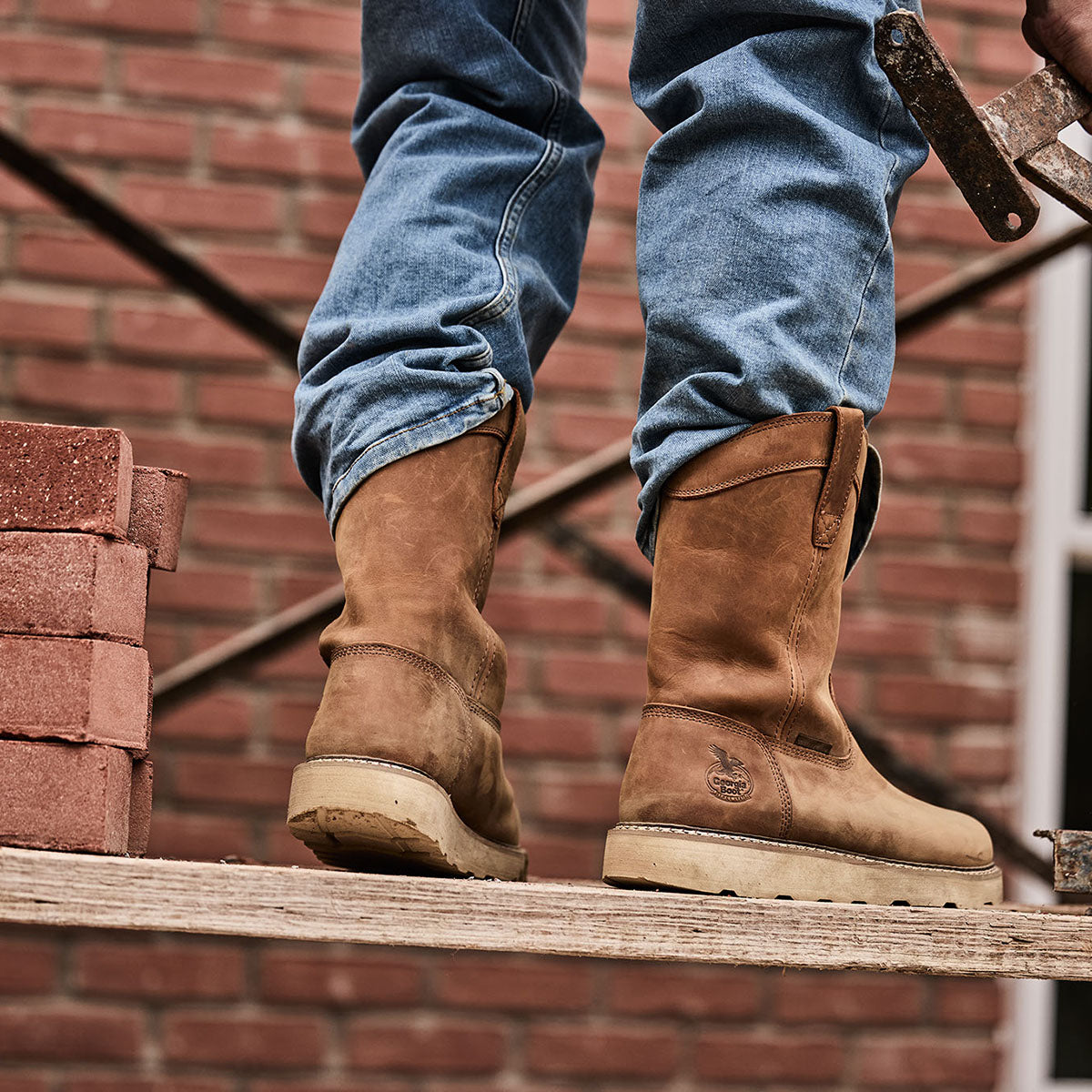 Person wearing brown work boots and blue jeans standing on a wooden platform with a brick wall background.