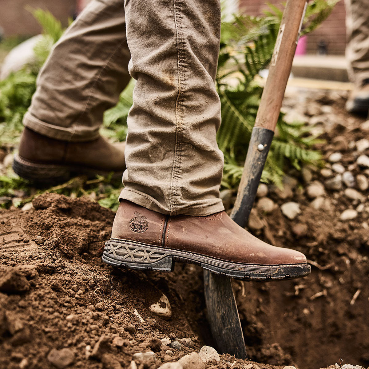Person wearing brown work boots and beige pants standing on a garden bed with a shovel.