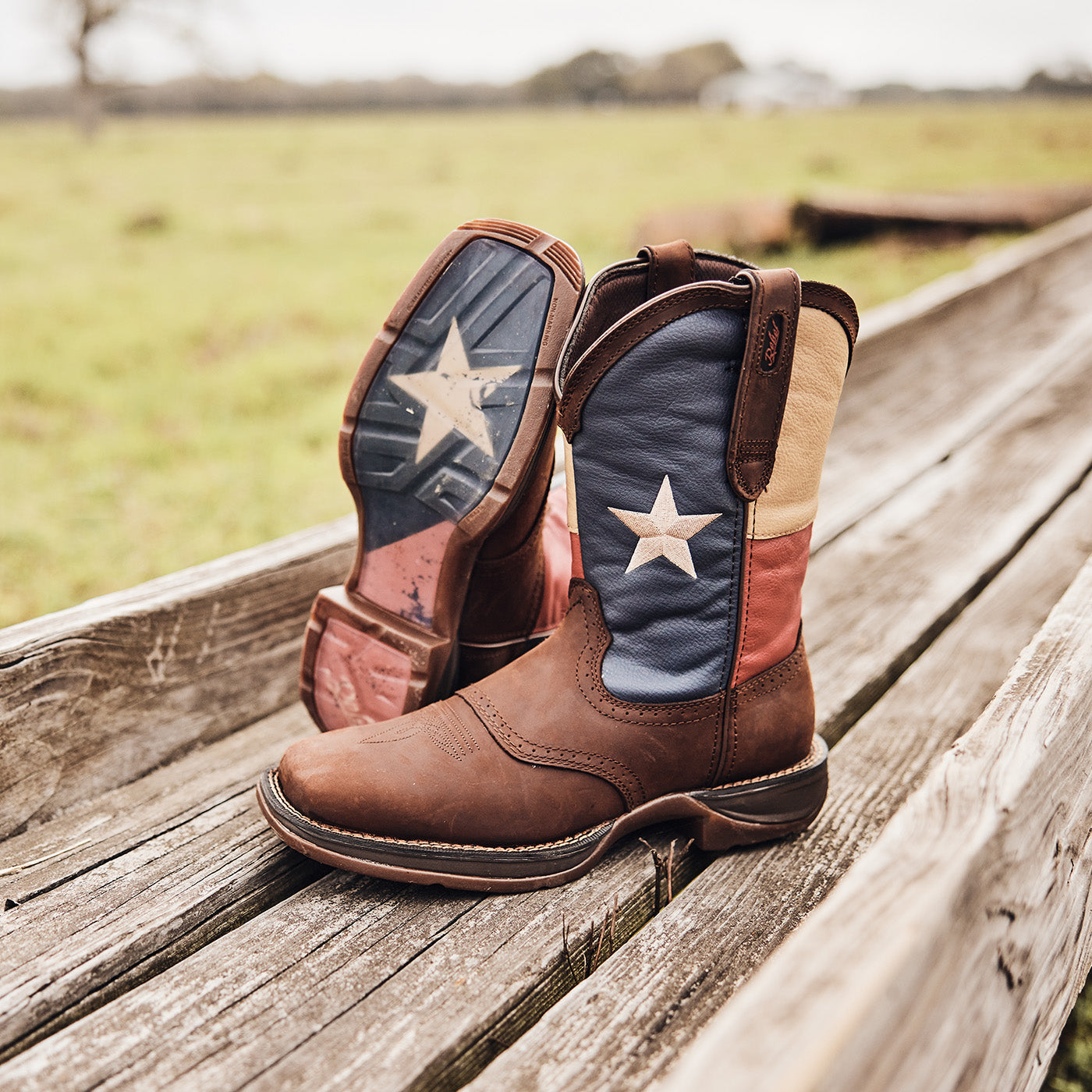 Brown cowboy boots with Texas flag pattern on a wooden surface with a grassy field background.