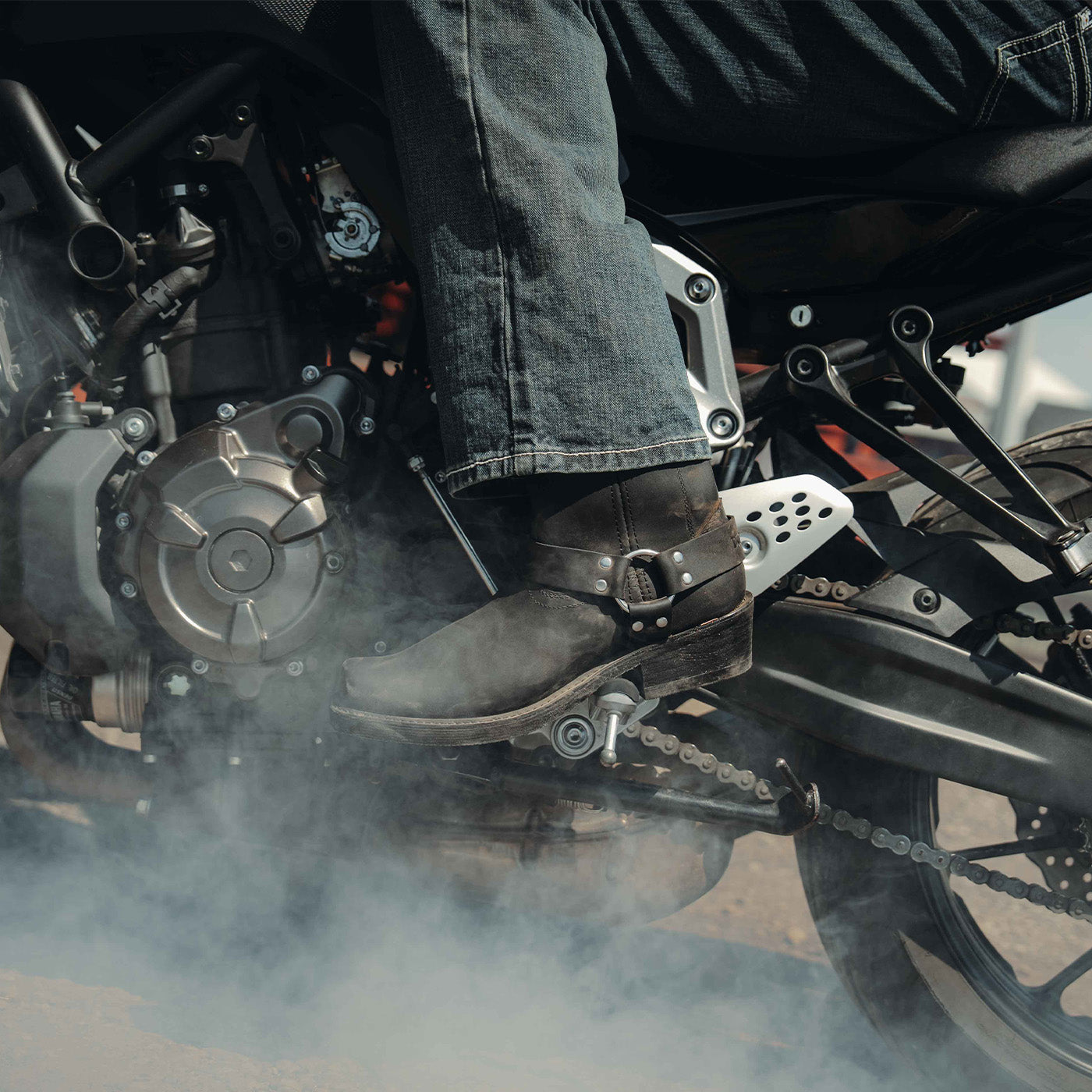 Close-up of a person's foot wearing a black Durango moto boot on a motorcycle with smoke in the background