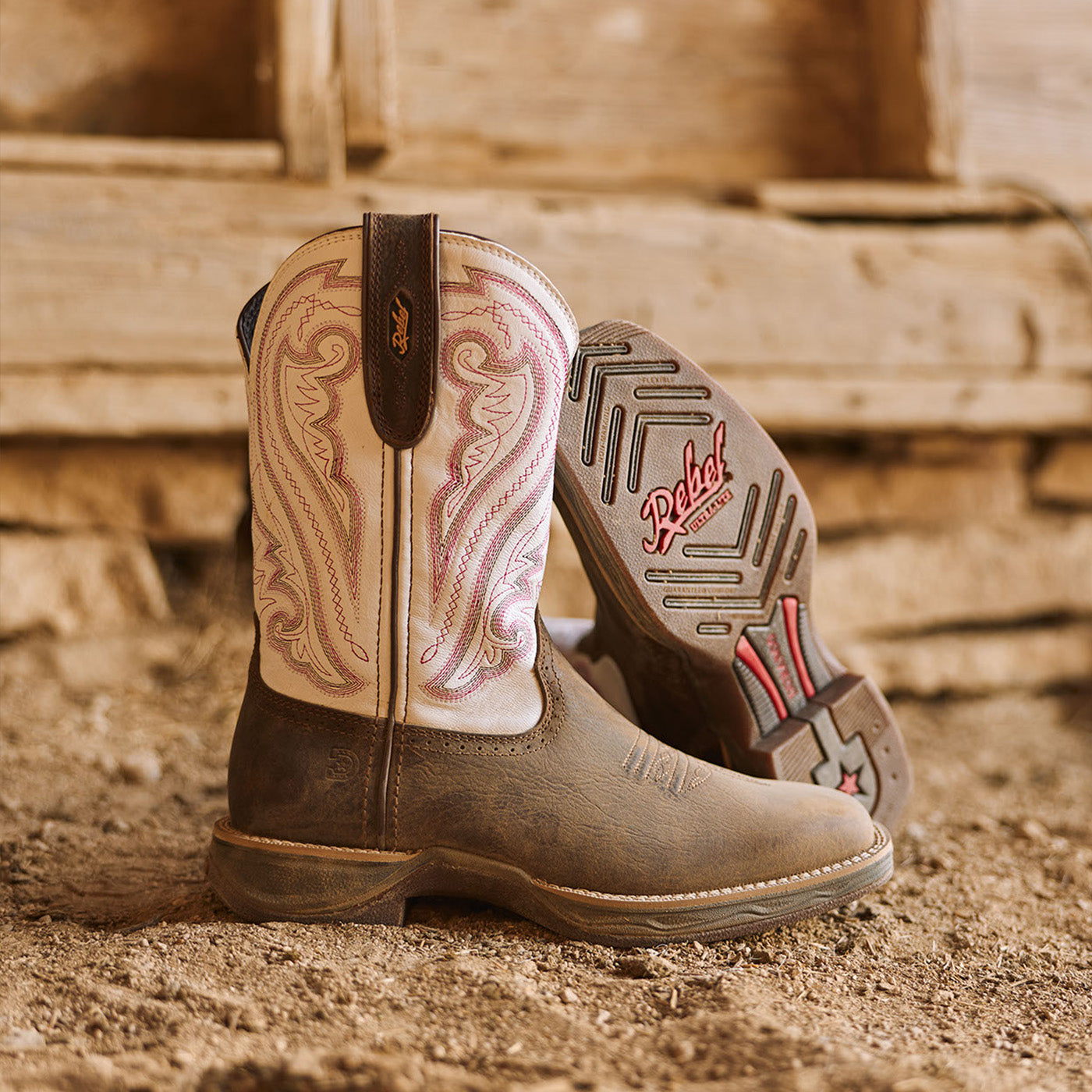 Brown and white women's Durango Rebel Ultra Lite western boots with pink stitching on a barn's dirt floor